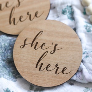 May include: Two wooden signs with the text "he's here" and "she's here" in cursive script. The signs are on a white and blue patterned fabric.