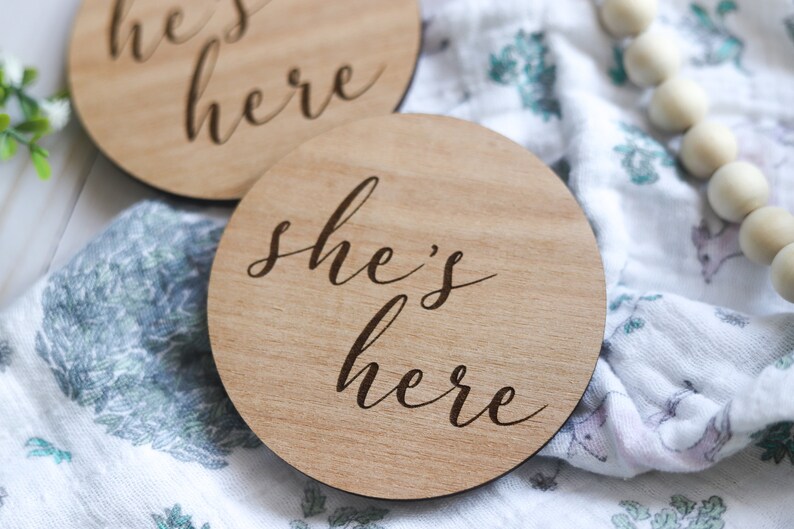 May include: Two wooden signs with the text "he's here" and "she's here" in cursive script. The signs are on a white and blue patterned fabric.