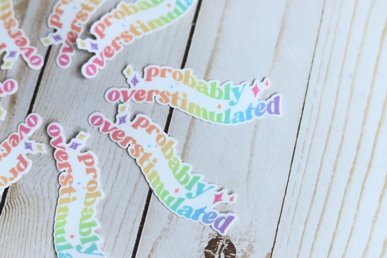 May include: A set of rainbow-colored stickers with the text "probably overstimulated" on each sticker. The stickers are on a light wood surface.