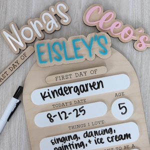 Pu&ograve; includere: Un cartello in legno per il primo giorno di scuola con il nome "Eisley's" in lettere blu. Il cartello include sezioni per "Kindergarten", data, et&agrave; e cose che amo. Sono visibili anche ritagli di nomi aggiuntivi.