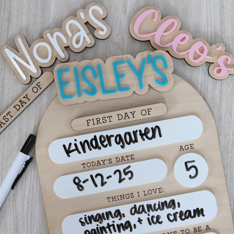 Pu&ograve; includere: Un cartello in legno per il primo giorno di scuola con il nome "Eisley's" in lettere blu. Il cartello include sezioni per "Kindergarten", data, et&agrave; e cose che amo. Sono visibili anche ritagli di nomi aggiuntivi.