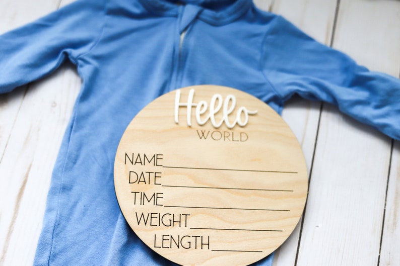 Op de afbeelding: Een rond houten bord met de tekst "Hello World" in witte letters. Het bord heeft lege regels voor "Naam", "Datum", "Tijd", "Gewicht" en "Lengte". Een blauwe baby romper ligt op een wit houten oppervlak.