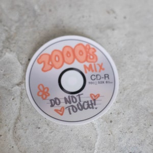 May include: A CD-R disc with the words "2000's MIX" in orange bubble letters. The disc also has the text "DO NOT TOUCH!" and a small flower and hearts. The disc is silver with a black center.
