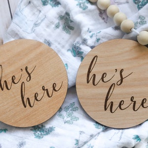 May include: Two wooden round signs with the text "she's here" and "he's here" in a cursive font. The signs are on a white and green patterned fabric.