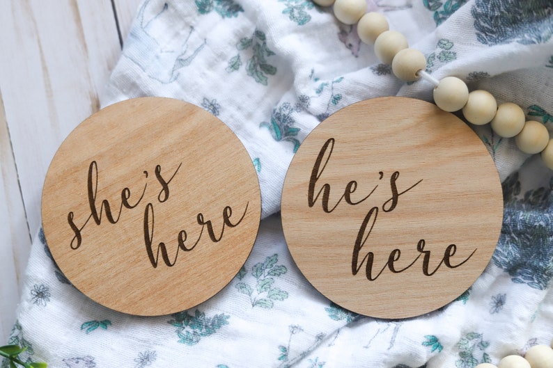 May include: Two wooden round signs with the text "she's here" and "he's here" in a cursive font. The signs are on a white and green patterned fabric.