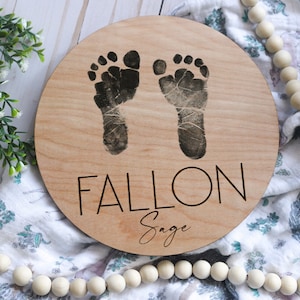 May include: A round wooden sign with two dark footprints and the name "FALLON" engraved in a bold font. The name "Sage" is written below in a cursive font. The sign is a light brown color.