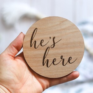 May include: A round wooden sign with the text "he's here" written in cursive.