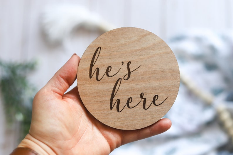 May include: A round wooden sign with the text "he's here" written in cursive.