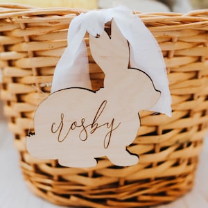 May include: A wooden bunny-shaped tag with the name "Crosby" engraved on it. The tag is attached to a wicker basket with a white ribbon.