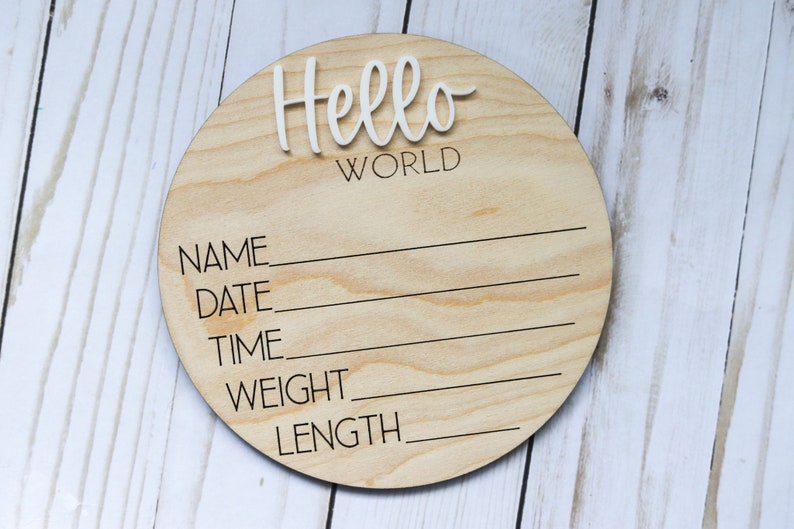 Op de afbeelding: Een rond houten bord met de tekst "Hello World" bovenaan. Onder de tekst staan lege regels voor "Naam", "Datum", "Tijd", "Gewicht" en "Lengte".