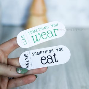 May include: Two white oval stickers with black and green text. The top sticker says "Cleo Something You Wear". The bottom sticker says "Kelsie Something You Eat".