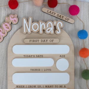 Pu&ograve; includere: Una lavagna di legno per i ricordi scolastici con il nome "Nora's" in lettere bianche. La lavagna include sezioni per "Today's Date", "Age" e "Things I Love". Un piccolo cartello di legno recita "Last Day Of". Pompon colorati sono infilati sopra.