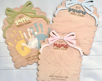 May include: Three scalloped wooden plaques with bows. One features a yellow and blue handprint and text reading Best Guppie Hands Down! Happy Mother's Day! Love, Nora, Cleo. Others read Best Grandma Hands Down! and Best Mama Hands Down! Happy Mother's Day! Nora, Cleo & Willa.