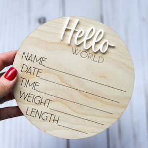 Op de afbeelding: Een rond houten bord met de woorden "Hello World" in witte letters. Het bord heeft lege regels om de naam, datum, tijd, gewicht en lengte van de baby in te vullen.