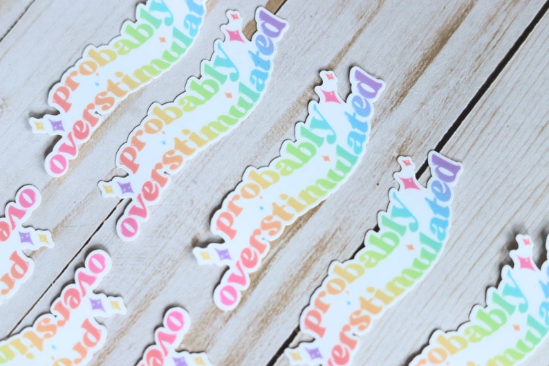 May include: A set of rainbow-colored stickers with the text "Probably Overstimulated" in a wavy font. The stickers are on a light wood background.