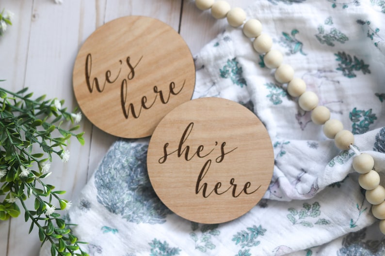 May include: Two wooden round signs with the text "he's here" and "she's here" in cursive script. The signs are on a white and blue patterned fabric with a string of wooden beads.