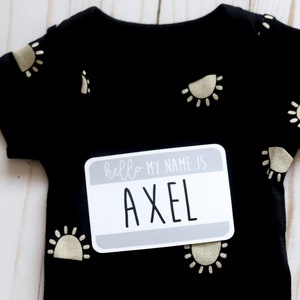 May include: Black baby onesie with gold sun print and a gray name tag that says "hello my name is AXEL".