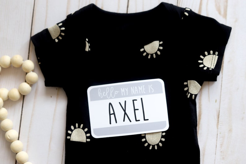 May include: Black baby onesie with gold sun print and a gray name tag that says "hello my name is AXEL".