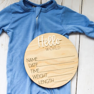 Op de afbeelding: Een rond houten bord met de tekst "Hello World" in witte letters. Het bord heeft lege regels voor "Name", "Date", "Time", "Weight" en "Length". Een blauwe baby romper ligt op een wit houten oppervlak.