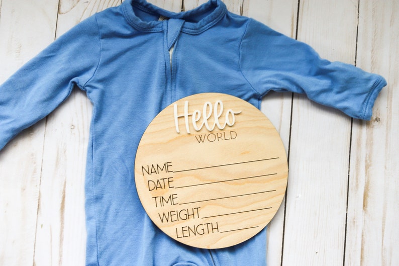 Op de afbeelding: Een rond houten bord met de tekst "Hello World" in witte letters. Het bord heeft lege regels voor "Name", "Date", "Time", "Weight" en "Length". Een blauwe baby romper ligt op een wit houten oppervlak.