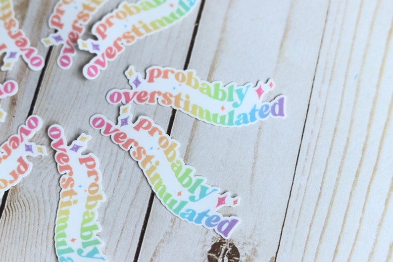 May include: A set of rainbow-colored stickers with the text "probably overstimulated" on each sticker. The stickers are on a light wood background.