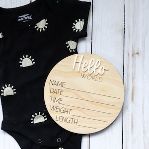 Op de afbeelding: Een rond houten bord met de tekst "Hello World" en lege regels voor naam, datum, tijd, gewicht en lengte. Het bord ligt naast een zwarte romper met witte zonontwerpen.