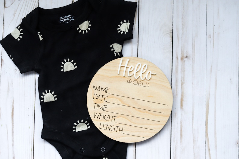 Op de afbeelding: Een rond houten bord met de tekst "Hello World" en lege regels voor naam, datum, tijd, gewicht en lengte. Het bord ligt naast een zwarte romper met witte zonontwerpen.