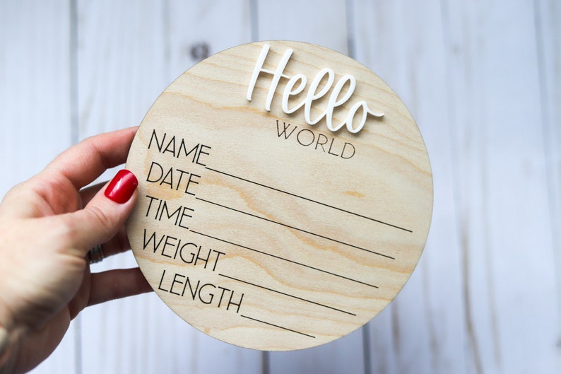 Op de afbeelding: Een rond houten bord met de woorden "Hello World" in witte letters. Het bord heeft lege regels om de naam, datum, tijd, gewicht en lengte van de baby in te vullen.