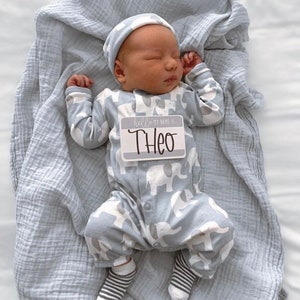 May include: A newborn baby wearing a gray and white elephant print onesie, a gray knit hat, and striped socks. The baby is holding a name tag that says "Hello my name is Theo".