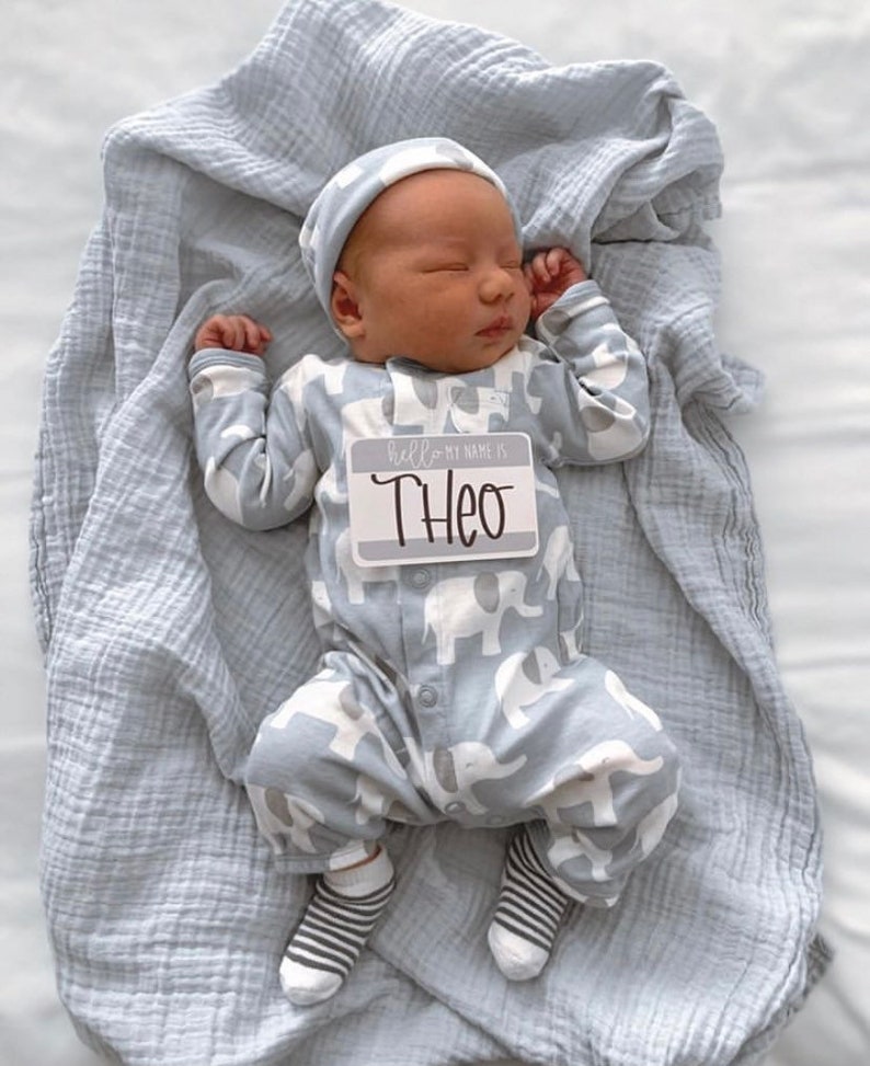 May include: A newborn baby wearing a gray and white elephant print onesie, a gray knit hat, and striped socks. The baby is holding a name tag that says "Hello my name is Theo".