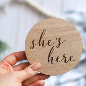 May include: A round wooden sign with the text "she's here" engraved in a cursive font.