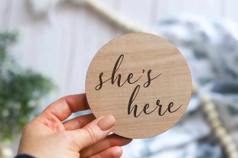 May include: A round wooden sign with the text "she's here" engraved in a cursive font.