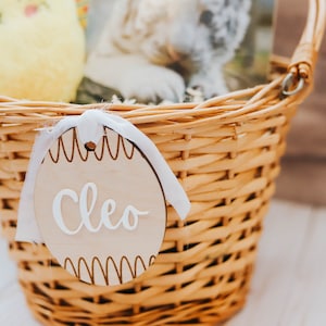 May include: A wicker basket filled with Easter treats, including a yellow chick toy and a book. A wooden tag with the name "Cleo" is attached to the basket.