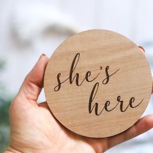 May include: A round wooden sign with the text "she's here" in a cursive font.