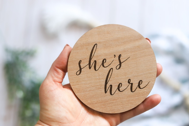 May include: A round wooden sign with the text "she's here" in a cursive font.