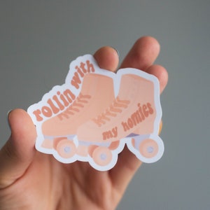 May include: A sticker featuring a pair of peach-colored roller skates with the words "rollin with my homies" in a matching peach color. The sticker is held in a hand against a gray background.
