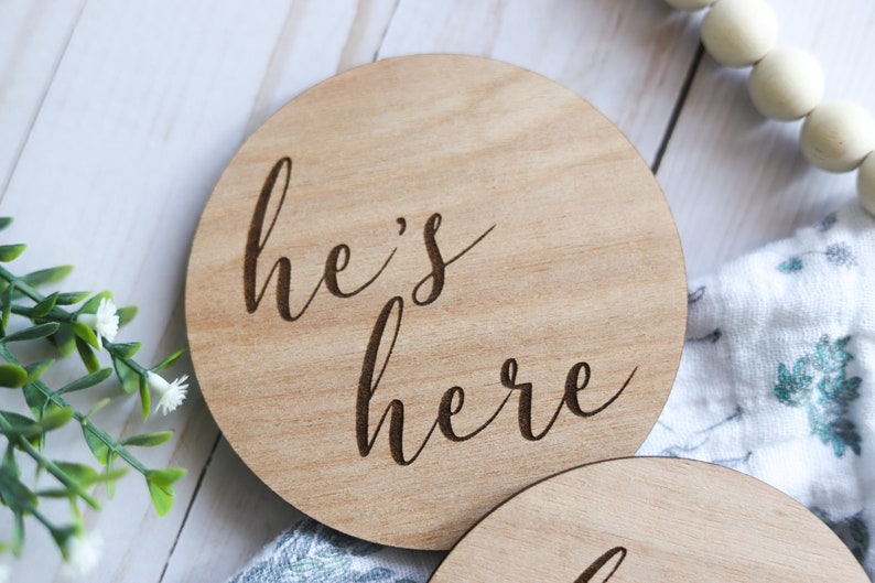May include: A round wooden sign with the text "he's here" engraved in a cursive font.
