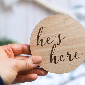 May include: A round wooden sign with the text "he's here" written in cursive.