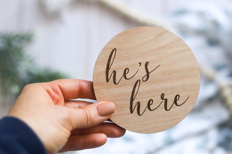 May include: A round wooden sign with the text "he's here" written in cursive.