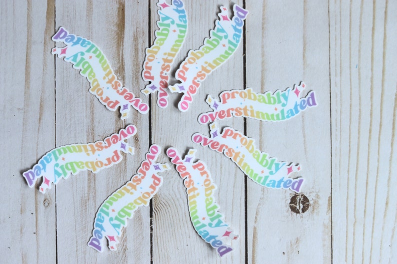 May include: A set of nine rainbow-colored stickers with the text "probably overstimulated" on each sticker. The stickers are arranged in a circular pattern on a light wood background.