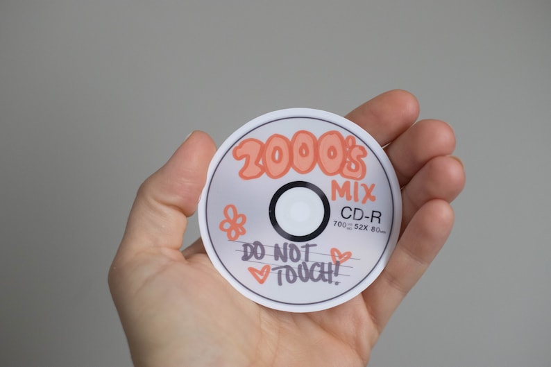 May include: A round sticker resembling a CD-R, held in a hand. The sticker has the text "2000's MIX" in orange, along with a flower and the words "DO NOT TOUCH!" in black. The CD-R has the dimensions 700MB, 52x80mm.