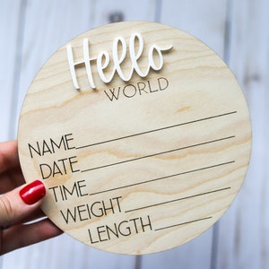 Op de afbeelding: Een rond houten bord met de tekst "Hello World" in witte letters. Het bord heeft lege regels voor "Naam", "Datum", "Tijd", "Gewicht" en "Lengte".