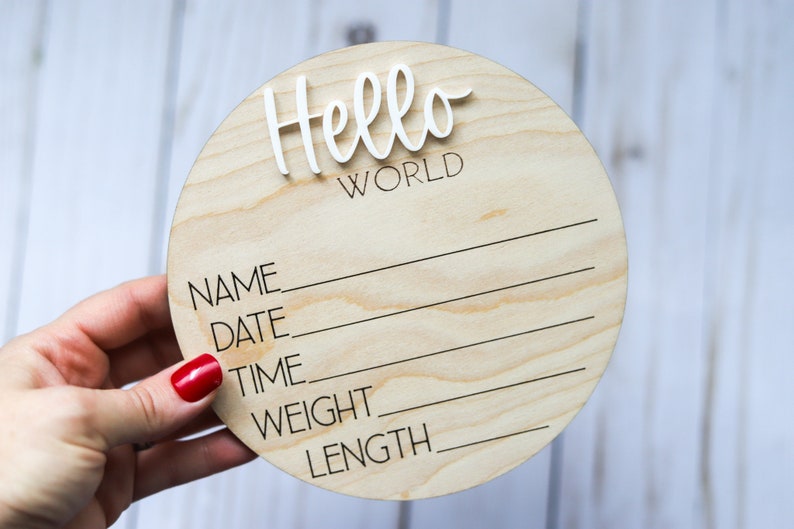Op de afbeelding: Een rond houten bord met de tekst "Hello World" in witte letters. Het bord heeft lege regels voor "Naam", "Datum", "Tijd", "Gewicht" en "Lengte".