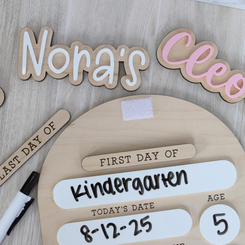 Pu&ograve; includere: Un cartello in legno per il primo giorno di scuola con le parole "First Day of Kindergarten" e "Today's Date 8-12-25". Include l'et&agrave; "5" e i nomi "Nora's" e "Cleo" in bianco e rosa.