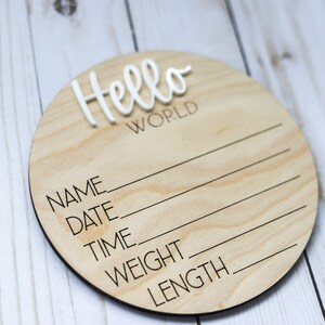 Op de afbeelding: Een rond houten bord met de tekst "Hello World" in witte letters. Het bord heeft lege regels voor "Naam", "Datum", "Tijd", "Gewicht" en "Lengte".