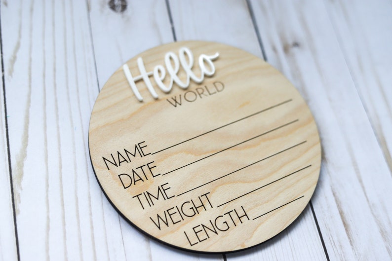 Op de afbeelding: Een rond houten bord met de tekst "Hello World" in witte letters. Het bord heeft lege regels voor "Naam", "Datum", "Tijd", "Gewicht" en "Lengte".