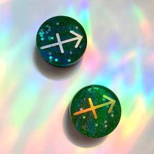 May include: Two green round resin ornaments with a gold and silver Sagittarius zodiac symbol. The ornaments are covered in glitter and have a green background.