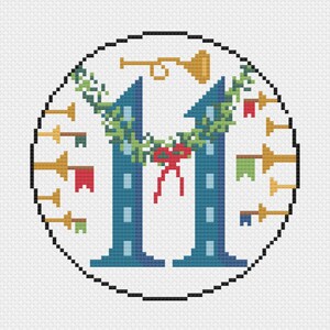Op de afbeelding: Een kruissteekontwerp met een blauwe letter "M" met een groene krans en een rode strik. Gouden trompetten en pijlen met kleurrijke vlaggen omringen de letter in een witte cirkel.