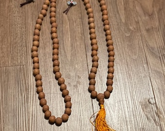 Puede incluir: Un collar de cuentas de mala de madera marrón con un borla de hilo naranja. El collar está colocado sobre una superficie de madera.
