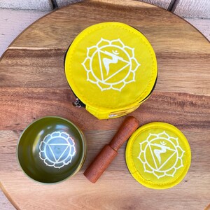 May include: A yellow singing bowl set with a wooden striker, a yellow pouch, and a yellow cushion. The bowl and cushion feature a white symbol of the solar plexus chakra.